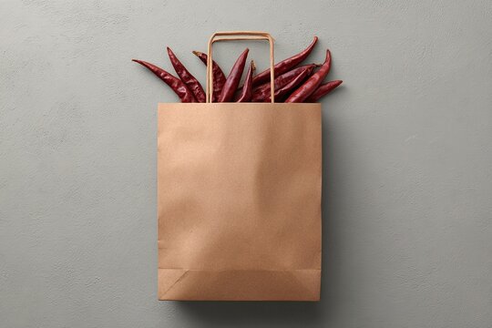 Brown paper shopping bag filled with dried red chili peppers on a textured gray background - Powered by Adobe