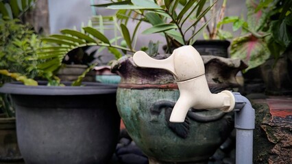 A white outdoor water tap attached to a pipe stands beside ceramic plant pots, surrounded by lush...