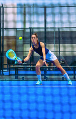 A focused female padel player stretches to reach an approaching ball during a match on a blue indoor court.