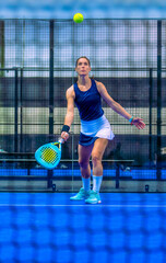 A determined female padel player looks up to hit the incoming ball on a blue indoor court.