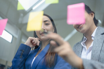 Two business woman with sticking adhesive notes on glass wall in office workplace, Creative startup with team concept.