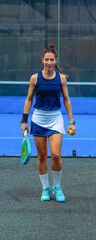 Smiling female padel player walking purposefully across the court holding the racket and ball, looking at and approaching the camera.
