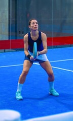 A focused, sharp, and active young woman holds her racket on a blue padel court, waiting for the ball with her eyes fixed on the court.