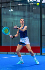 An athletic woman in sportswear and a padel skirt balances perfectly, waiting for the ball to come towards her from above during an indoor padel match.