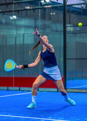 An athletic woman in blue sportswear and a padel skirt prepares herself in the perfect position for a groundstroke on an indoor padel court.