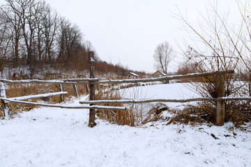 A rustic wooden fence encloses a snowy area near a frozen pond in the countryside. This quiet winter landscape with bare trees and small distant huts evokes a peaceful atmosphere during the cold seaso