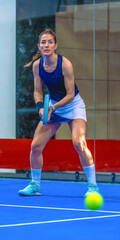 A determined, attentive, and confident female padel player in a ready position with the ball in the foreground approaching her on an indoor padel court
