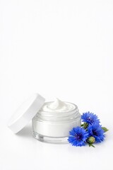 Glass jar of cream with white lid, near blue flowers. Beauty product. Remedy for face or skin around eyes with cornflower extract. Vertical banner, copy space to advertise cosmetics white background
