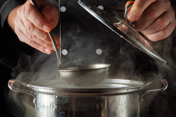 A chef lifts a lid from a pot filled with steaming soup. They hold a ladle in one hand and prepare to serve the hot dish. The kitchen is warm and busy during lunch