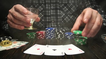 A man sits at a table with poker chips and playing cards. He holds a drink in one hand and tosses a green chip onto three aces with the other. The atmosphere is casual and lively