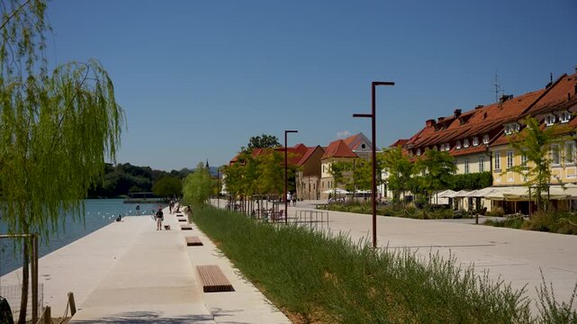 Maribor, Slovenia - August 9, 2025: A sunny riverside promenade in Maribor, Slovenia, features modern benches, young trees, and people relaxing by the water near traditional red-roofed buildings.