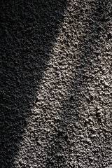 Rough stucco wall illuminated by sunlight casting diagonal grid shadows. The high-contrast light accentuates the deep granular texture of the concrete surface, creating a geometric abstract patter