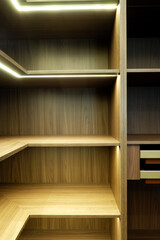Close-up of empty custom wooden wardrobe shelves featuring rich walnut grain texture. Recessed warm LED strip lighting under each shelf illuminates the corner joinery and dark timber finish.