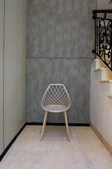 Modern white chair with a lattice backrest and wooden legs standing in a home corner. The background features a gray fluted wall panel and a marble staircase with a black wrought iron railing.