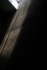 Vertical view of a rough textured wall illuminated by a diagonal beam of sunlight from a ceiling skylight. High contrast shadows create a dramatic industrial architectural pattern.