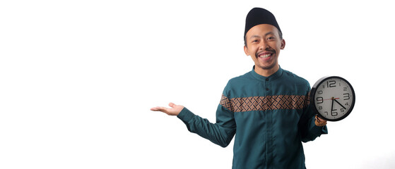 Asian man wearing a green traditional Muslim outfit, extending one hand to the side toward empty copy space while holding a wall clock in his other hand. Isolated on a white background