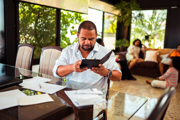 Focused man reviewing documents on a tablet at a glass table in a bright, modern home setting with family in the background.