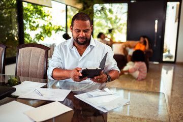 African American man working from home, reviewing documents and using a tablet at a glass desk in a bright, contemporary living space.