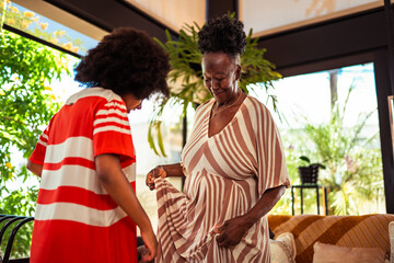 Smiling senior Black woman adjusts her striped dress while a young woman observes in a bright, plant-filled home.