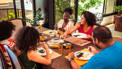 Obraz premium Happy multi-generational Black family enjoying a homemade meal together at a dining table, sharing food and laughter in a bright home setting.