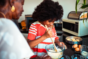 Happy African American girl bakes with her grandmother in the kitchen, mixing ingredients for cookies.