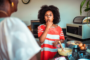 Cute Black girl tastes delicious batter from her finger while baking with an adult in a home kitchen.