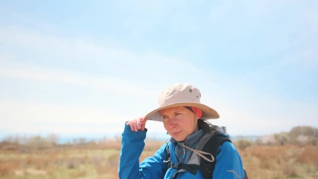 A woman wearing a wide-brimmed hat, blue hiking jacket, and backpack rides a horse through dry open grassland in sunny weather. She adjusts her hat while enjoying the outdoor adventure. 