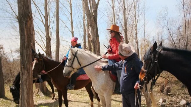 A group of women on a horseback ride take a sunny break in a leafless forest. Dressed warmly in hats and jackets, they enjoy the moment, gently petting their horses and relaxing with a guide nearby.