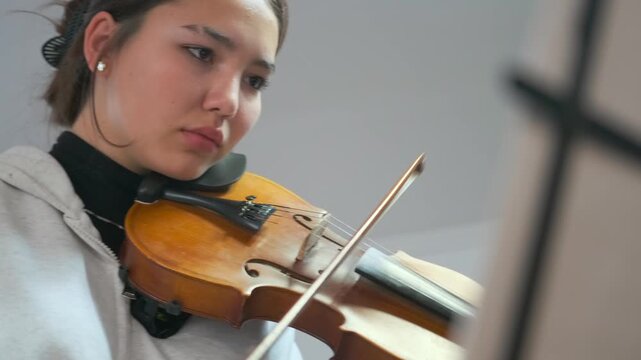 Close-up of a young female violin student carefully following sheet music while playing. Her focused expression, smooth bowing, and the classical instrument, home practice or private study.