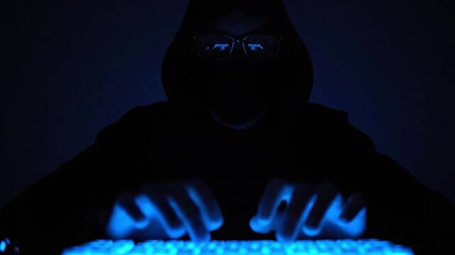A mysterious, silhouetted figure is shown typing intently on a vibrant, glowing blue keyboard in a dimly lit, high-tech environment. The individual, wearing a hooded garment and glasses, has their fac
