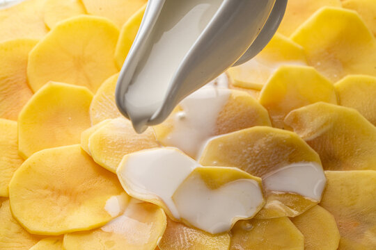 The process of making potato gratin. Delicate cream is poured over raw, thin potato slices.
