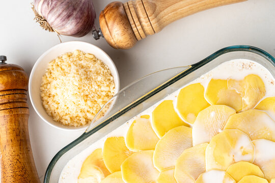 Preparation of potato gratin. Glass mold with raw potato slices with cream, cheese, spices and garlic on a white table.