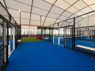 Spacious indoor padel court featuring a vivid blue surface, surrounded by metal fences and overhead...