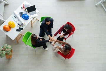 A diverse startup team stacks hands in unity during a planning session. The top view captures collaboration, innovation, and teamwork in a creative workspace with helmets, blueprints, and office tools