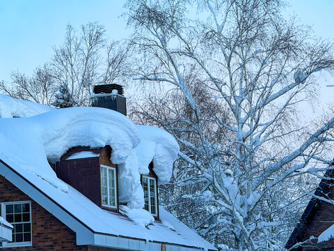 CLOSE UP: A thick blanket of icy snow covers the roof of a suburban red brick house on a cold winter day in Niseko, Japan. Snow-laden roof arcs over windows, with birch trees fully covered in snow.