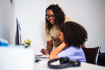 African American mother helping her young daughter with homework or online learning at a desk in a home office.