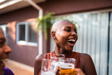 Happy young Black woman with shaved head laughing and clinking beer glasses with friends at an outdoor backyard party.