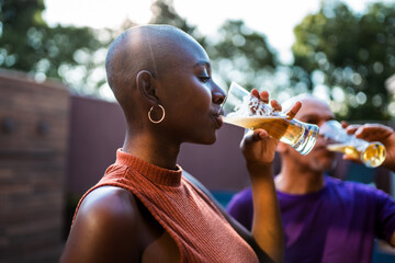 Young Black woman with shaved head drinking cold beer at an outdoor social gathering during sunset.