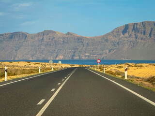 Tarmac road stretches out towards the Atlantic Ocean, with volcanic cliffs rising in the distance. A sunny, remote island road in a dry, vast landscape, ideal for road trips and coastal adventures.