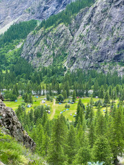 Picturesque mountain valley with a sprawling green campsite dotted with numerous tents and vehicles. Dense forests and steep mountain slopes surround campground in a natural, alpine setting in summer.