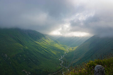 Scenic alpine valley stretches into the mist with a winding river carving through vibrant green meadows. Grey cloud cover drapes mountain slopes in a soft haze, serene atmosphere in the Swiss Alps.