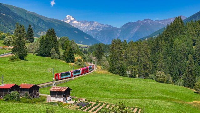 AERIAL: Train travels through a picturesque Swiss alpine valley, passing through pine forests, rustic villages and with views of majestic mountains. Scenic route through the mountainous countryside.