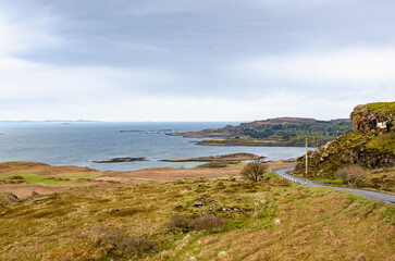 Coastal road on the Isle of Mull, Scotland, winding through grassy hills above a calm Atlantic bay under cloudy skies. Remote Scottish Highlands seascape with copy space.