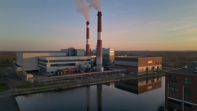 Industrial facility with smokestacks emitting white smoke beside reflective water at sunset in rural landscape