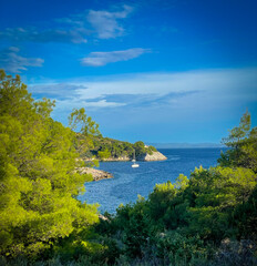 White sailboat rests in a sheltered turquoise bay surrounded by lush green pine forests. Bright sunlight and blue water create a peaceful Mediterranean scene perfect for a relaxing summer getaway.