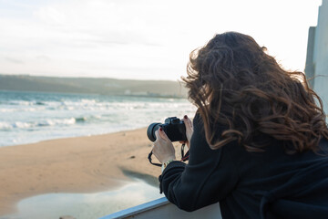 Smiling female photographer holding a camera on a sunny beach, enjoying golden hour light and capturing outdoor moments on the coastline.