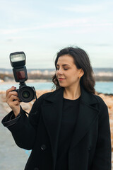 Young female photographer taking pictures outdoors by the sea, smiling while holding a professional camera with flash, capturing moments on a rocky coastline at sunset.