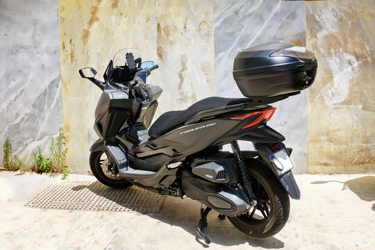 Bormla, Malta - July 13, 2025: A modern matte black Honda Forza 350 maxi-scooter with a top box parked against a decorative marble wall on a bright sunny day