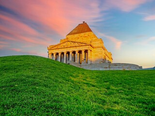 Fototapeta premium Shrine of Remembrance in Melbourne, Australia, glowing in the warm light of sunset. The historic war memorial sits atop a grassy hill against a dramatic pink and blue sky.