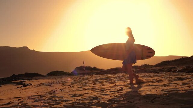 LENS FLARE, SILHOUETTE: Woman in skort and with surfboard in hands is walking towards the beach when she stops to check the waves. She is going for sunset surf session at beautiful Lanzarote island.